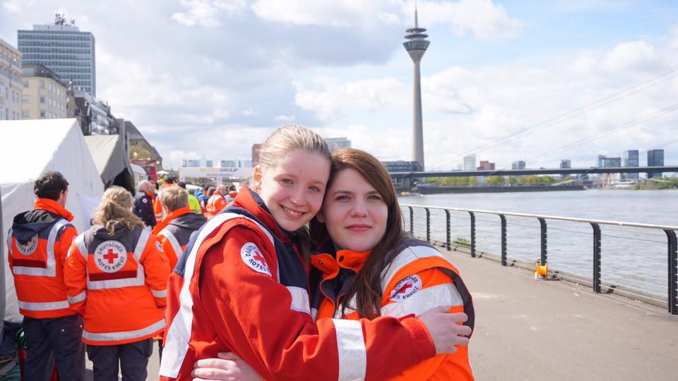 zwei Rettungssanitäterinnen umarmen sich am Rhein. Im Hintergrund ist der Rheinturm zu sehen.