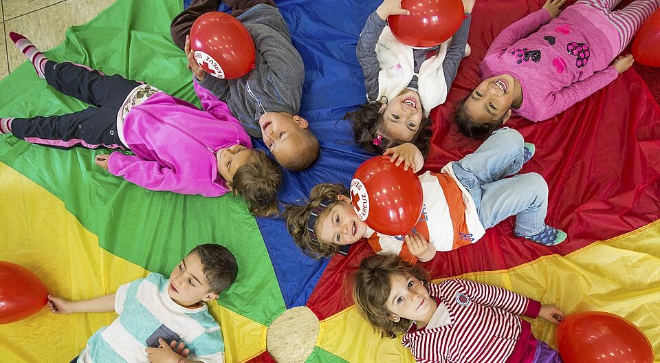 lachende Kinder liegen auf buntem Tuch mit DRK Luftballons