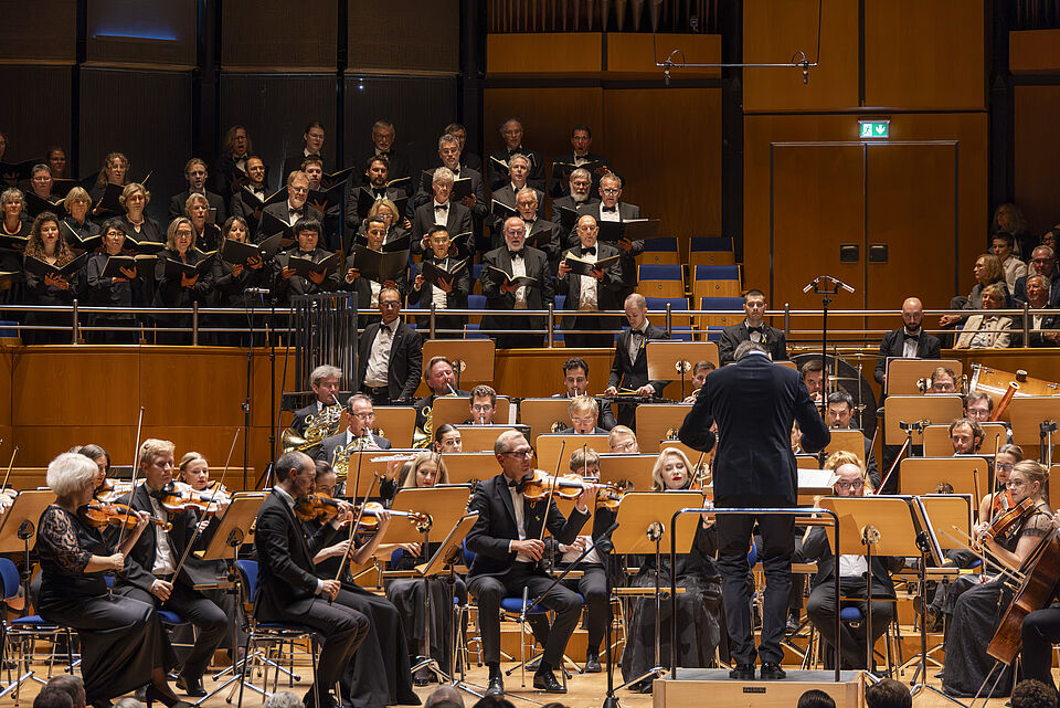 Blick aufs Orchester auf der Bühne und darüber im Rang ein Chor